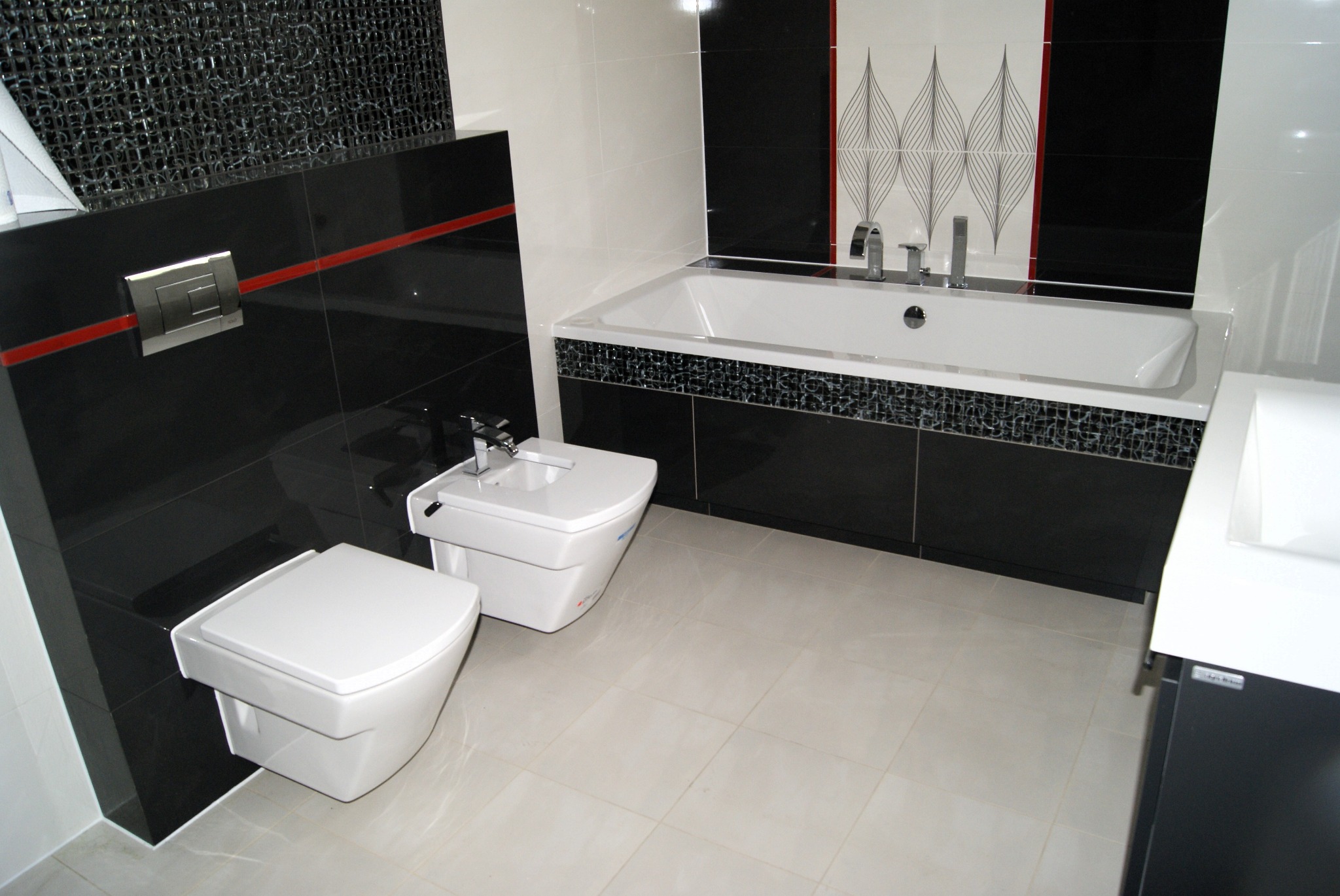 Nowoczesna łazienka z białymi sanitariatami wiszącymi na tle czarnych płytek z czerwoną linią dekoracyjną, mozaiką nad wanną i wzorzystą tapetą.