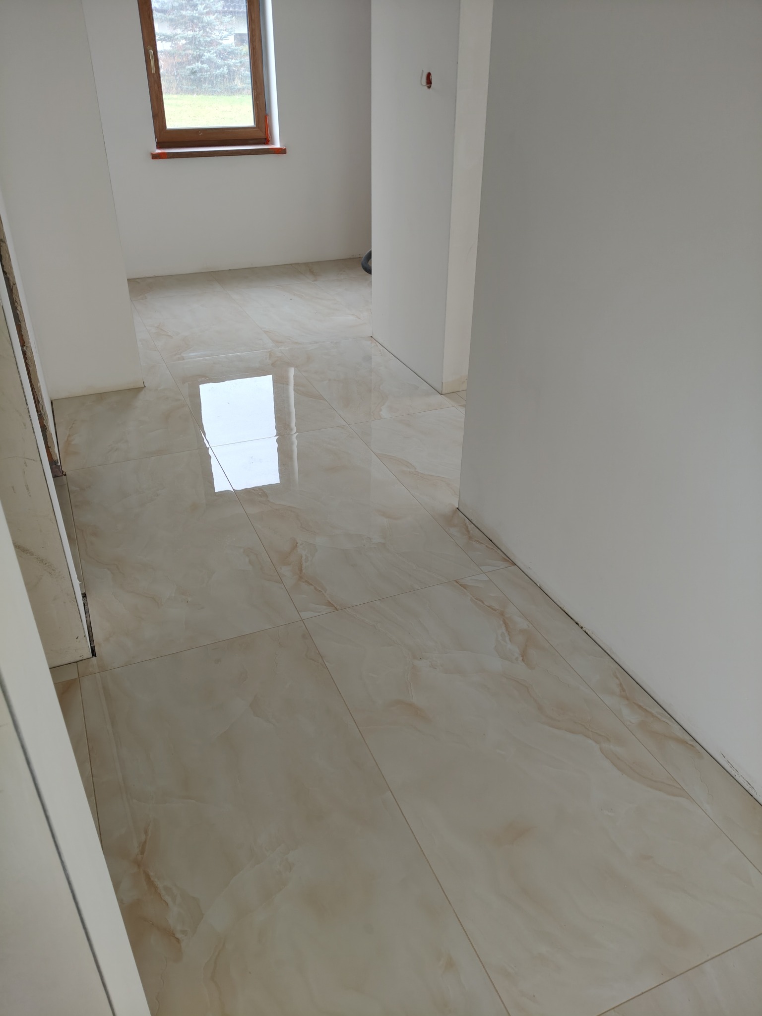 Połyskująca, beżowa podłoga z płytek imitujących marmur w jasnym wnętrzu. Widoczne fragmenty ścian i okno z drewnianą ramą. Minimalistyczny styl.