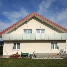 Kam-Weld - Jasny dom jednorodzinny z balkonem zabezpieczonym mlecznymi panelami, na trawniku wózek ogrodowy, drewno i zabawki dziecięce, błękitne niebo z chmurami w tle.