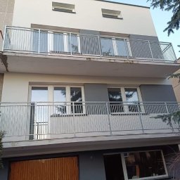 Kam-Weld - Dwupiętrowy budynek z balkonami zabezpieczonymi metalowymi balustradami o minimalistycznym designie, widok z dołu, z fragmentem zieleni drzew po prawej stronie.