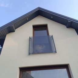 Kam-Weld - Szklana balustrada balkonowa z ciemnymi, metalowymi elementami mocującymi, widok z dołu na elewację budynku w jasnym kolorze z ciemnym dachem i oknami.