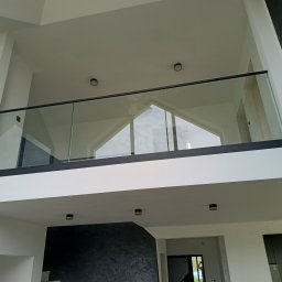 Kam-Weld - Wnętrze domu z antresolą zabezpieczoną szklaną balustradą z czarnymi profilami, widok z dołu na sufit z oświetleniem punktowym.