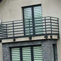 Kam-Weld - Czarna metalowa balustrada balkonowa z poziomymi szczeblami na budynku z szarą elewacją i oknami z żaluzjami.