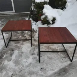 Dwa stoliki kawowe z drewnianymi blatami i metalowymi, czarnymi nogami, ustawione na zaśnieżonym podłożu przed budynkiem i krzewem.