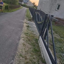 Kam-Weld - Metalowa brama z ozdobnym wzorem w kształcie serc, zamontowana na betonowej podmurówce, widoczna z perspektywy ulicy w wiejskiej scenerii z biegnącym mężczyzną w tle.