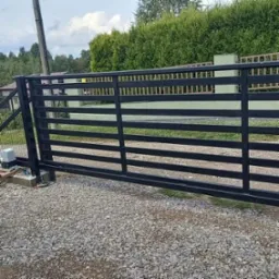 Czarna, przesuwna brama wjazdowa z poziomymi szczeblami, widoczny napęd elektryczny, podłoże z szarego żwiru, tło z zielonych krzewów i drzew.