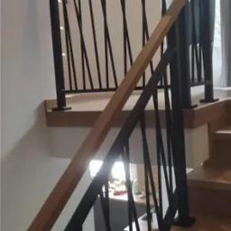 Wnętrze domu ze schodami o drewnianych stopniach i czarnej, metalowej balustradzie o geometrycznym wzorze, widok z dołu.