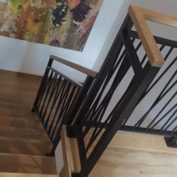 Kam-Weld - Nowoczesna balustrada schodowa z czarnymi, ukośnymi prętami i drewnianym pochwytem, widok z góry na drewniane schody i abstrakcyjny obraz na ścianie.