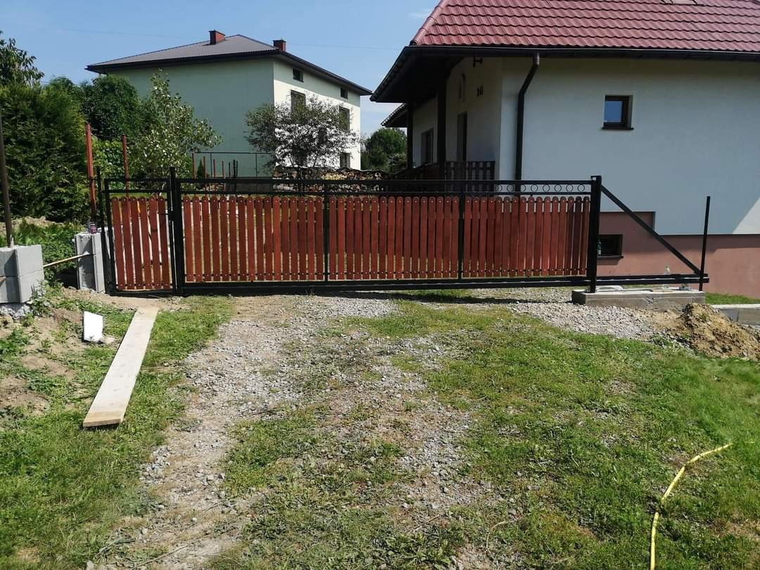 Suwana brama wjazdowa z drewnianymi sztachetami w kolorze brązowym i czarną metalową ramą, widoczna na tle dwóch domów jednorodzinnych w słoneczny dzień.