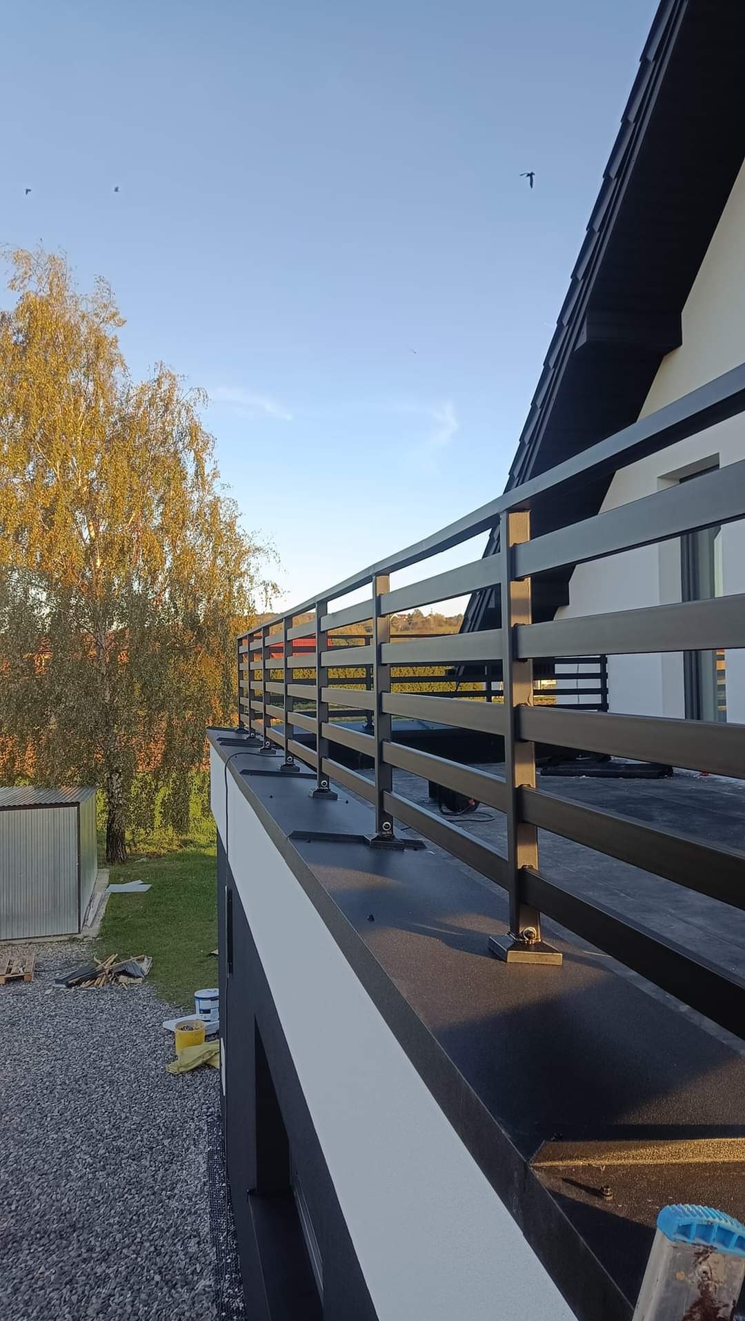 Czarna, metalowa balustrada balkonowa z poziomymi szczeblami, zamontowana na płaskim dachu nowoczesnego domu, widok z boku na tle jesiennego drzewa i błękitnego nieba.