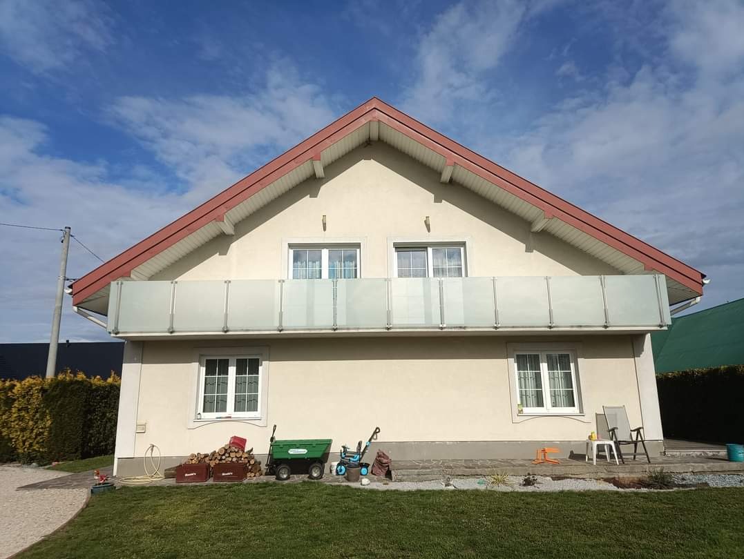 Jasny dom jednorodzinny z balkonem zabezpieczonym mlecznymi panelami, na trawniku wózek ogrodowy, drewno i zabawki dziecięce, błękitne niebo z chmurami w tle.