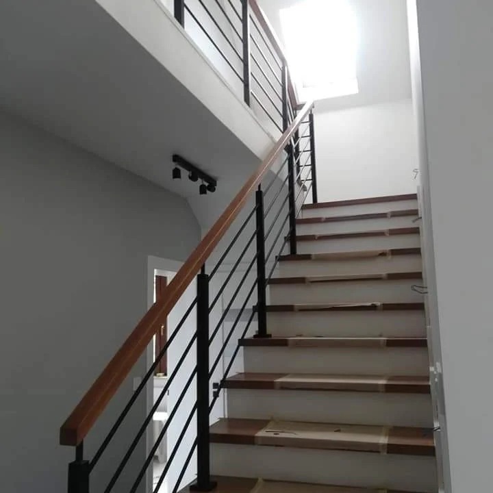 Wnętrze domu: schody z drewnianymi stopniami zabezpieczonymi folią, czarne metalowe barierki i drewniana poręcz, widok z dołu, oświetlenie sufitowe.