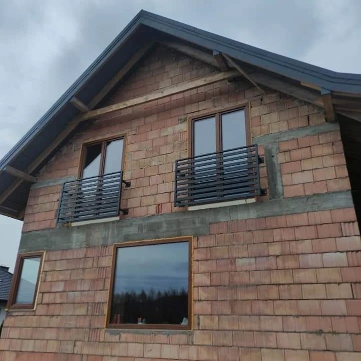 Surowy stan budynku z cegły z zamontowanymi, czarnymi, metalowymi balustradami balkonowymi przy oknach na piętrze. Widoczne niedokończone obróbki murarskie.