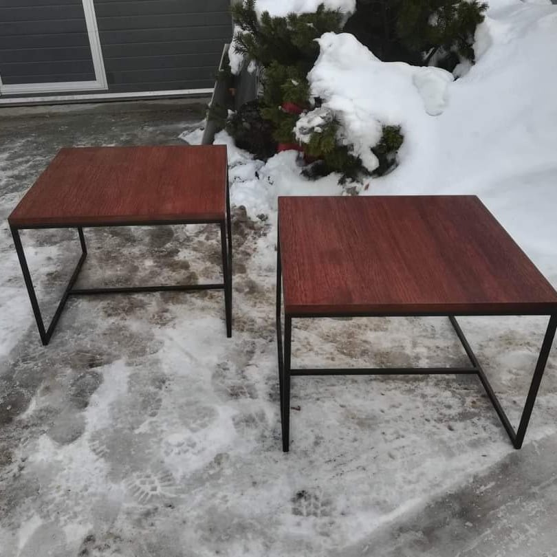 Dwa stoliki kawowe z drewnianymi blatami i metalowymi, czarnymi nogami, ustawione na zaśnieżonym podłożu przed budynkiem i krzewem.