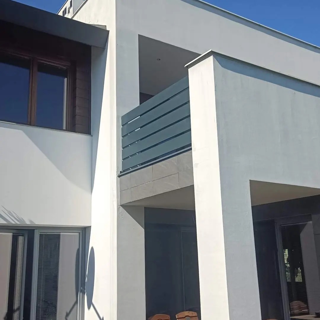 Nowoczesny dom z balkonem zabezpieczonym poziomą balustradą z szarych profili, widok z dołu na elewację z dużymi oknami.
