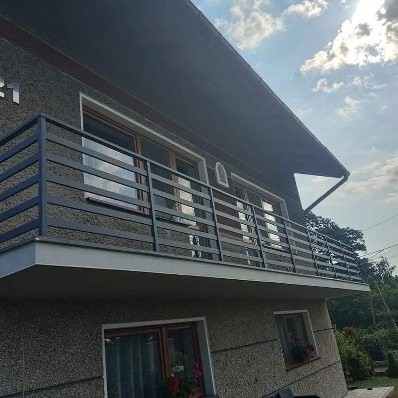 Szary dom z balkonem zabezpieczonym nowoczesną, metalową balustradą z poziomych profili, widok z dołu na tle błękitnego nieba z chmurami.