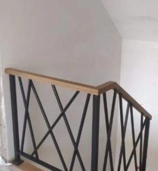 Wewnętrzna balustrada schodowa z czarnych, krzyżujących się profili stalowych i drewnianym pochwytem, zamontowana przy białej ścianie.