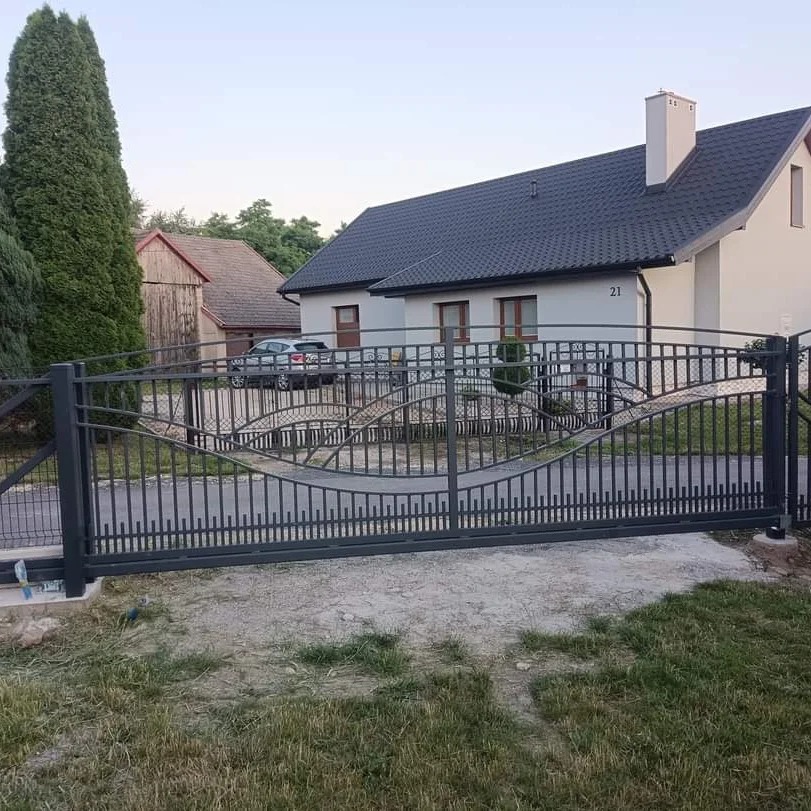 Nowoczesna, przesuwna brama wjazdowa z metalowymi prętami o falistym wzorze na tle domu z ciemnym dachem.