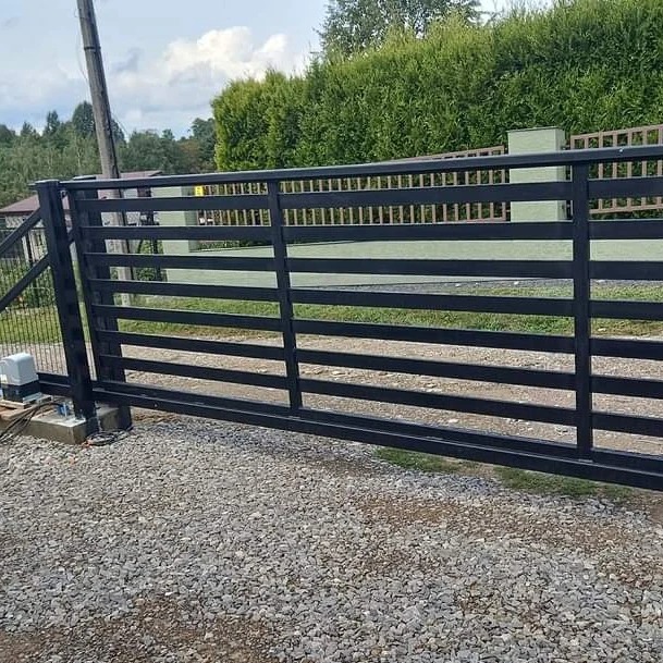 Czarna, przesuwna brama wjazdowa z poziomymi szczeblami, widoczny napęd elektryczny, podłoże z szarego żwiru, tło z zielonych krzewów i drzew.