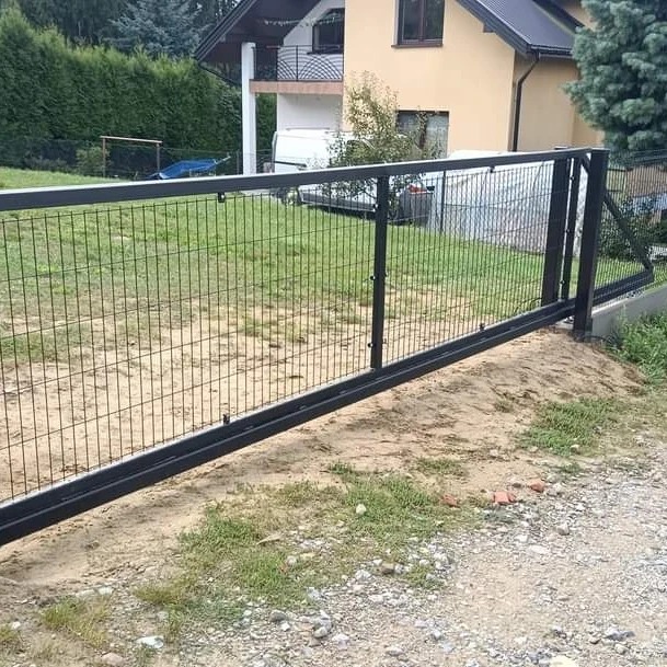 Przesuwna brama wjazdowa z czarnej siatki metalowej przed domem z balkonem; widoczny teren przygotowany pod kostkę brukową.