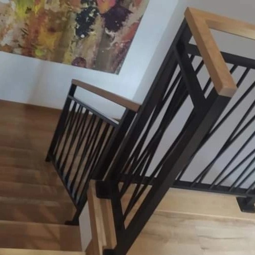 Nowoczesna balustrada schodowa z czarnymi, ukośnymi prętami i drewnianym pochwytem, widok z góry na drewniane schody i abstrakcyjny obraz na ścianie.