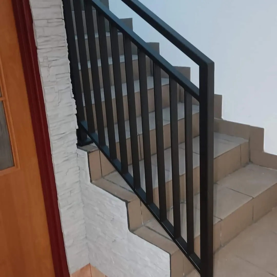 Schody wewnętrzne z czarną, metalową balustradą o prostym wzorze, widok z dołu schodów, z fragmentem drzwi po lewej stronie.