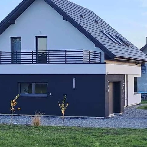 Nowoczesny dom z balkonem zabezpieczonym czarną, metalową balustradą, ciemnoszara elewacja, jasne okna, dachówka w kolorze grafitowym.