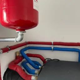 Czerwony zbiornik wyrównawczy ciśnienia w instalacji grzewczej, połączony srebrnymi rurami i czerwono-niebieskimi rurami izolowanymi, widoczne narzędzie zaciskarka do rur.