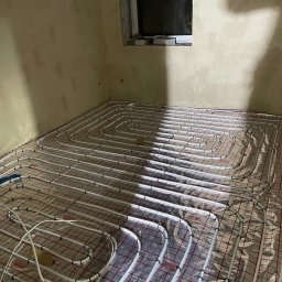 Melek-Instal - Rozłożona instalacja ogrzewania podłogowego na folii z siatką w surowym pomieszczeniu, widoczne białe rury i niebieski element mocujący.