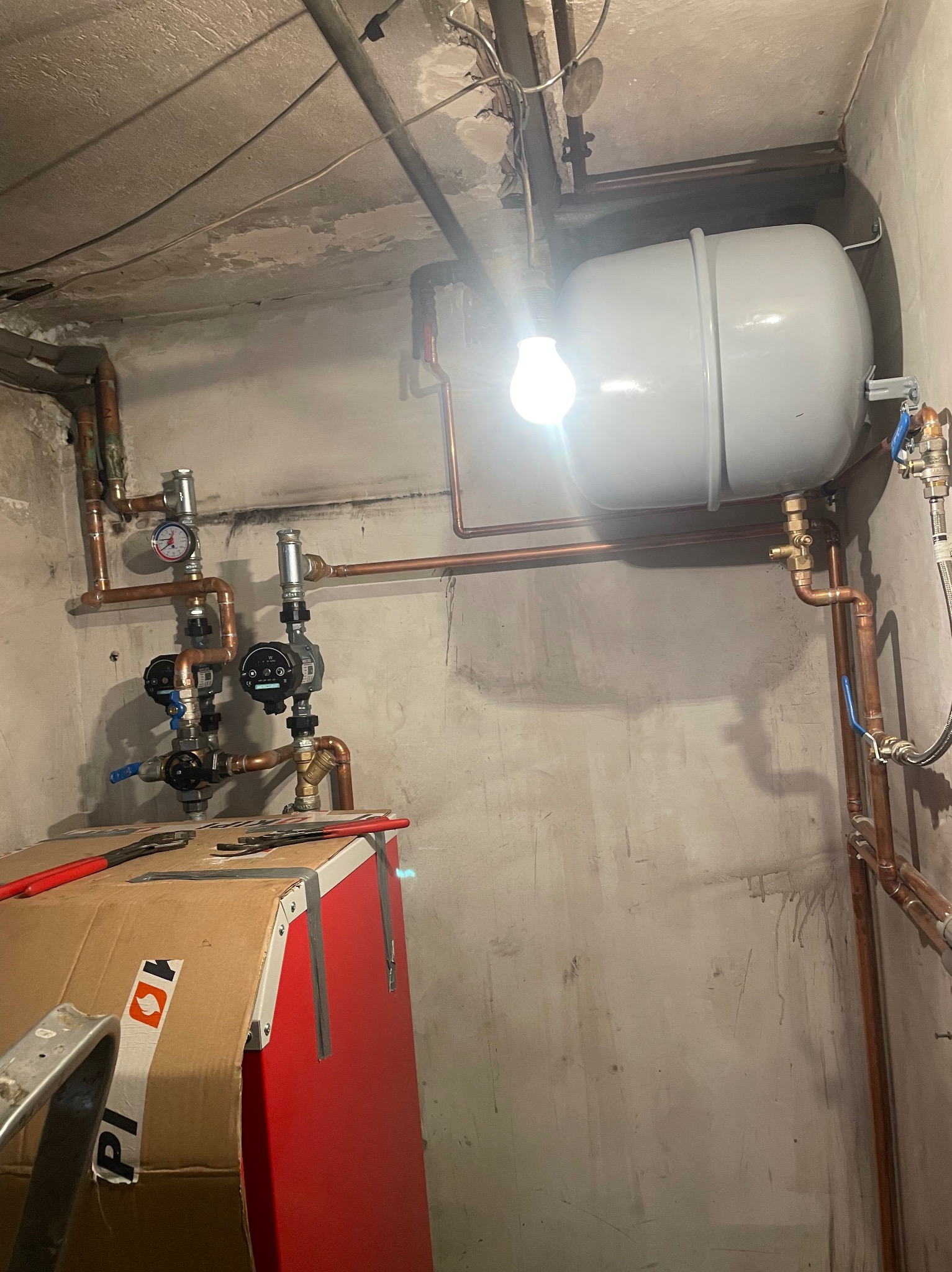 Montaż instalacji grzewczej w piwnicy: widoczny zbiornik wyrównawczy, pompy obiegowe, manometry, zawory oraz rury miedziane. Na pierwszym planie czerwony piec z kartonowym zabezpieczeniem...