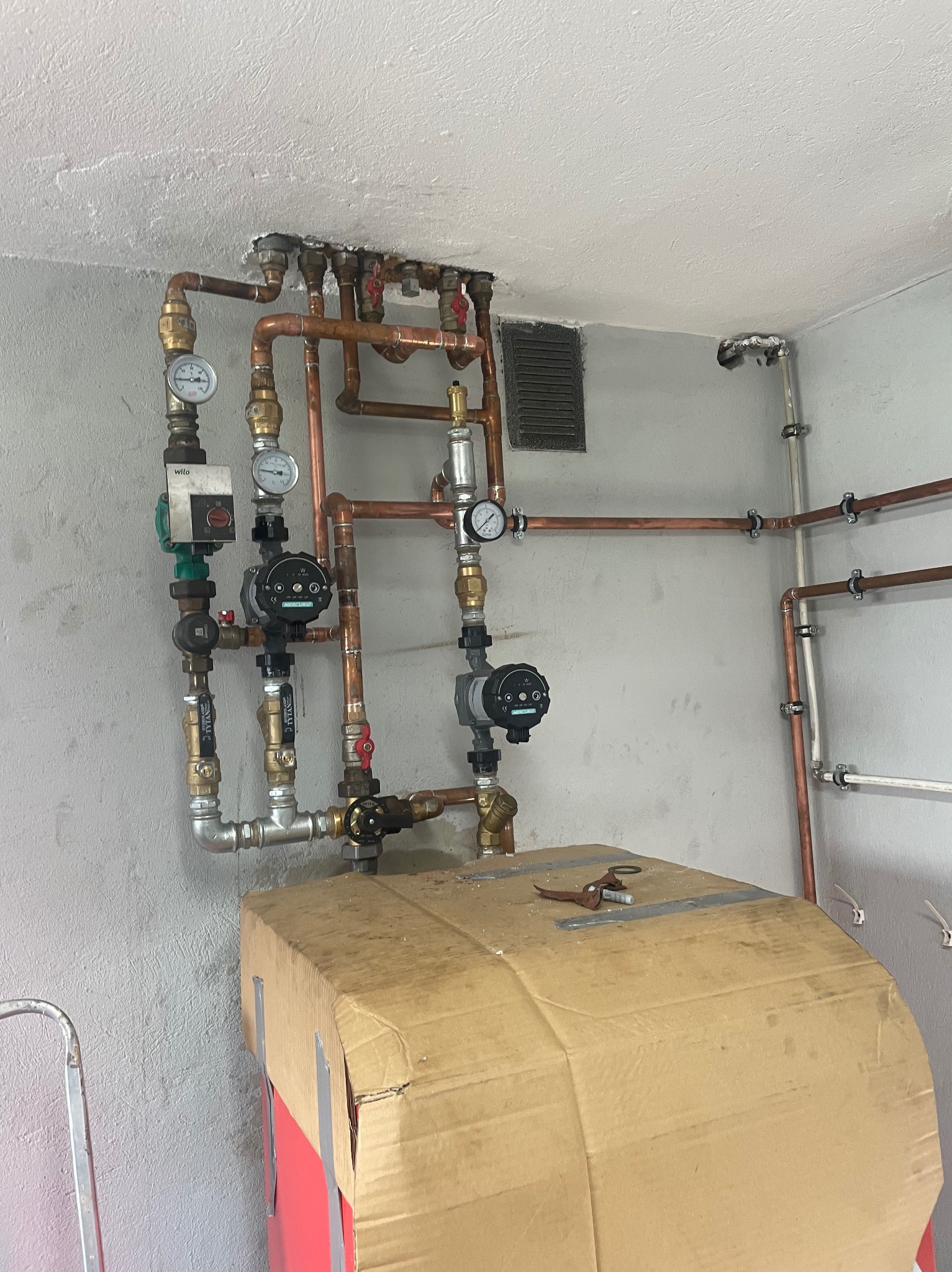 Rozbudowana instalacja grzewcza z miedzianymi rurami, manometrami i pompami obiegowymi, częściowo zasłonięta kartonem ochronnym, widoczne narzędzia na górze kartonu.