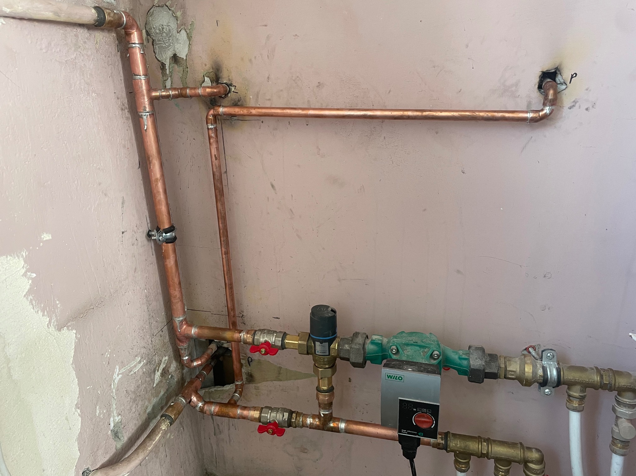 Miedziana instalacja centralnego ogrzewania z pompą obiegową Wilo, widoczne lutowane połączenia, zawory kulowe z czerwonymi rączkami, montaż na tle ściany z widocznymi śladami po kuciu.
