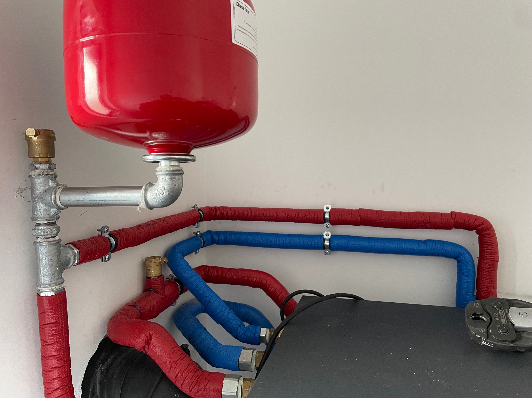 Czerwony zbiornik wyrównawczy ciśnienia w instalacji grzewczej, połączony srebrnymi rurami i czerwono-niebieskimi rurami izolowanymi, widoczne narzędzie zaciskarka do rur.