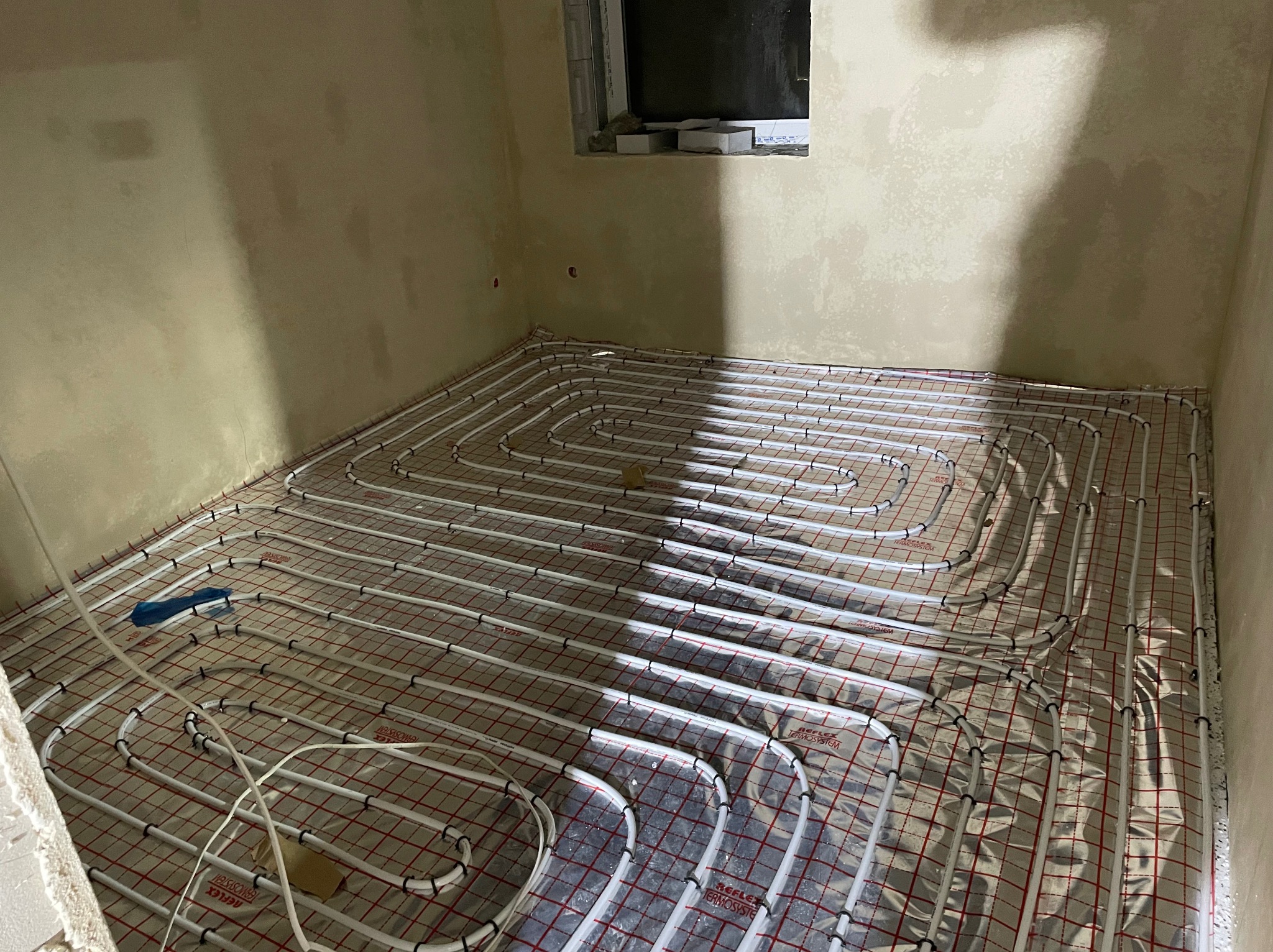 Rozłożona instalacja ogrzewania podłogowego na folii z siatką w surowym pomieszczeniu, widoczne białe rury i niebieski element mocujący.