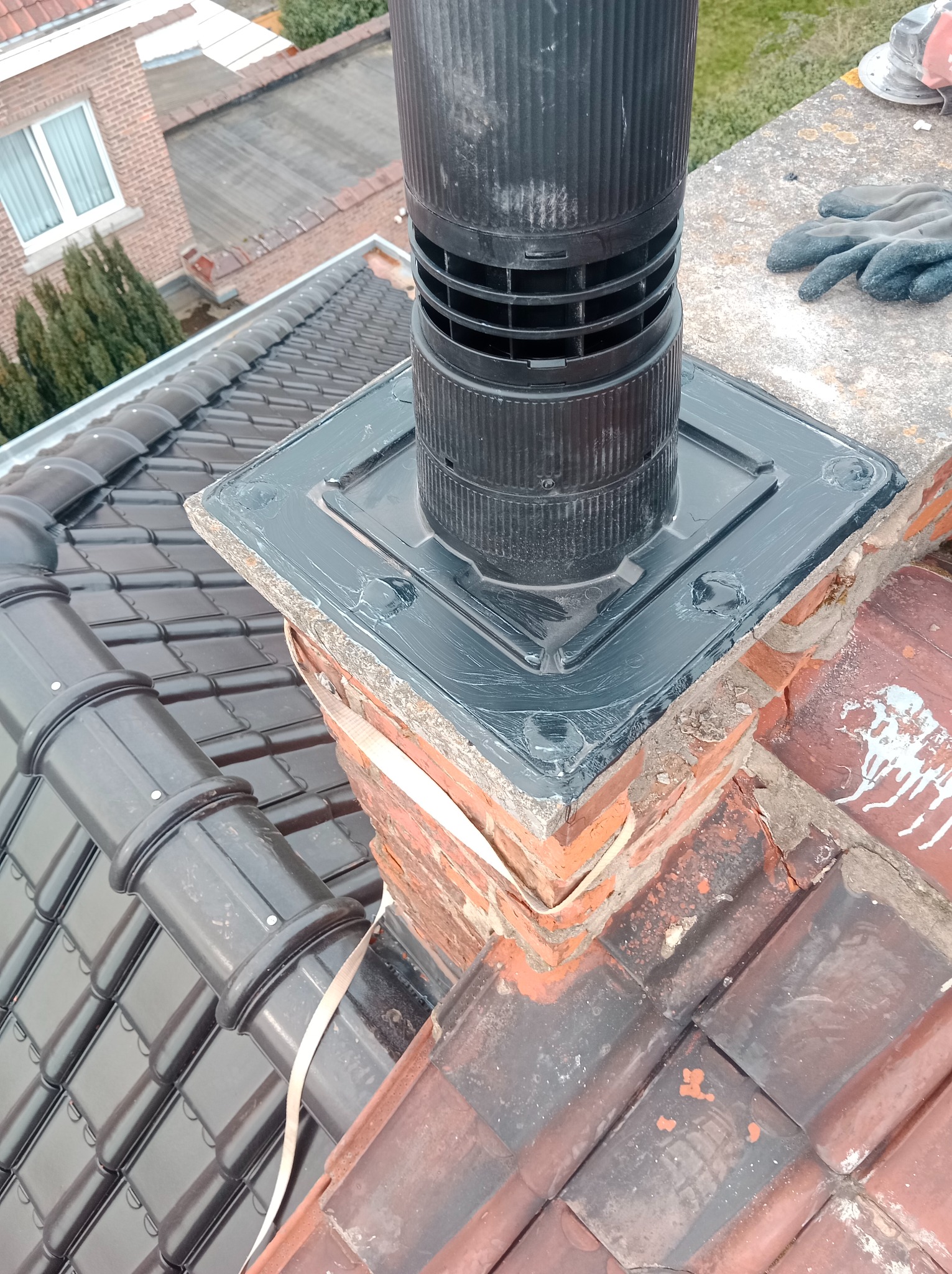 Czarny komin wentylacyjny zamontowany na ceglanym kominie na dachu pokrytym ciemną dachówką ceramiczną. Widoczne ślady uszczelniania silikonem wokół podstawy komina i robocze rękawice na dachu.