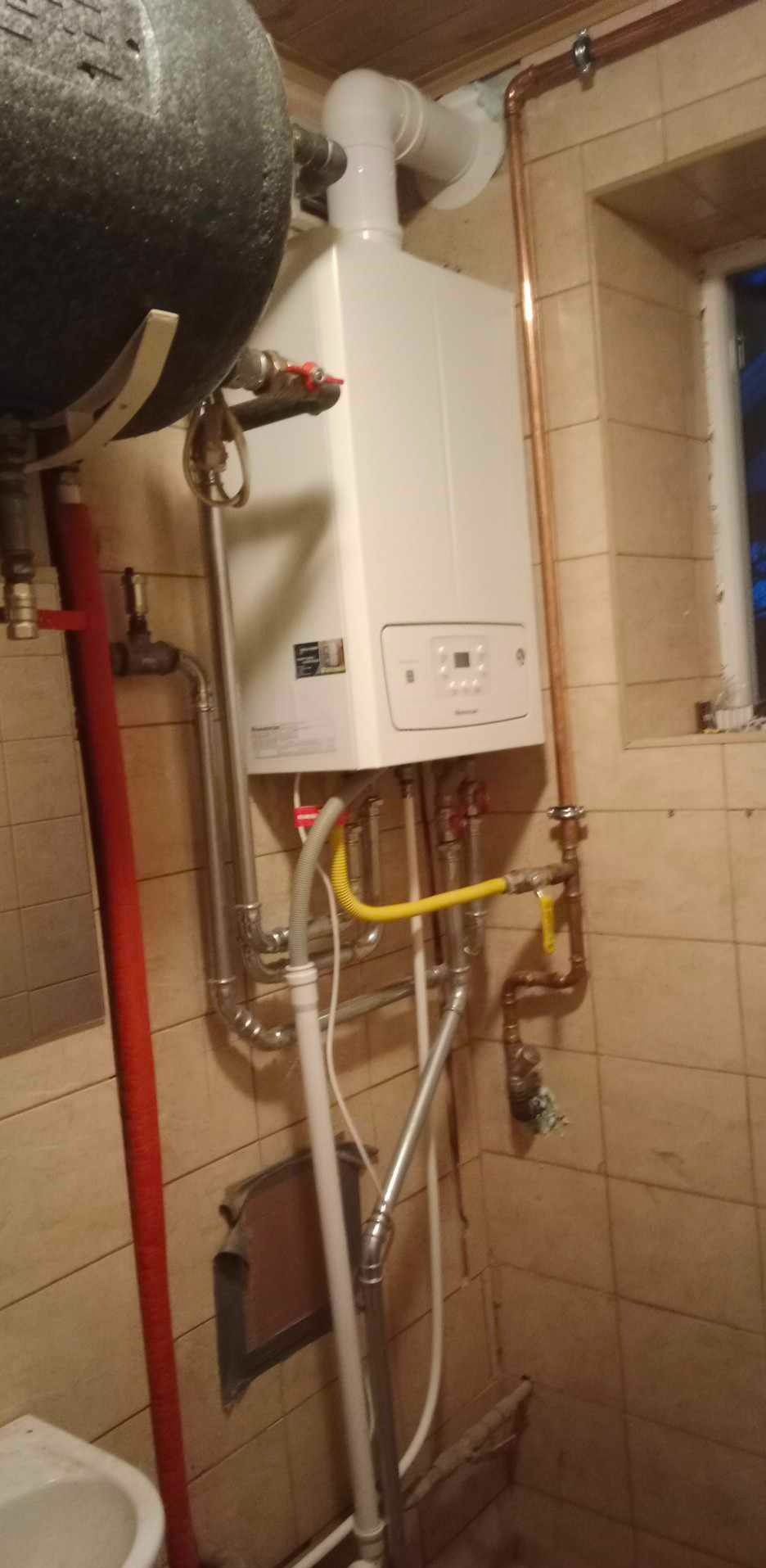 Biały piec CO z podłączeniami w Ługowiskach, widoczne rury miedziane i żółty przewód gazowy na tle beżowych płytek.