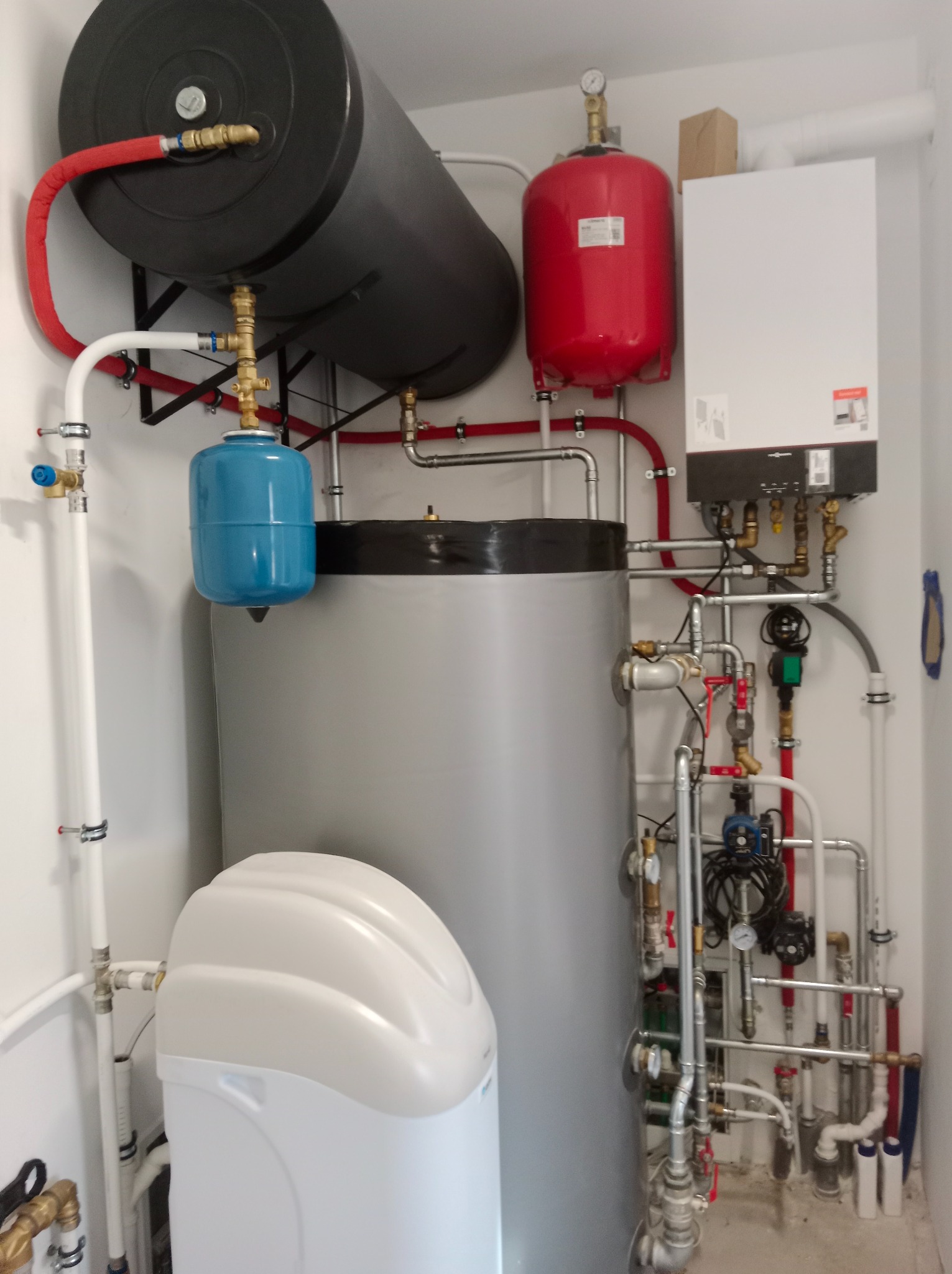 Kompletna instalacja kotłowni w pomieszczeniu: zasobnik ciepłej wody użytkowej, zbiornik wyrównawczy, piec grzewczy oraz stacja uzdatniania wody z licznymi rurami i zaworami.