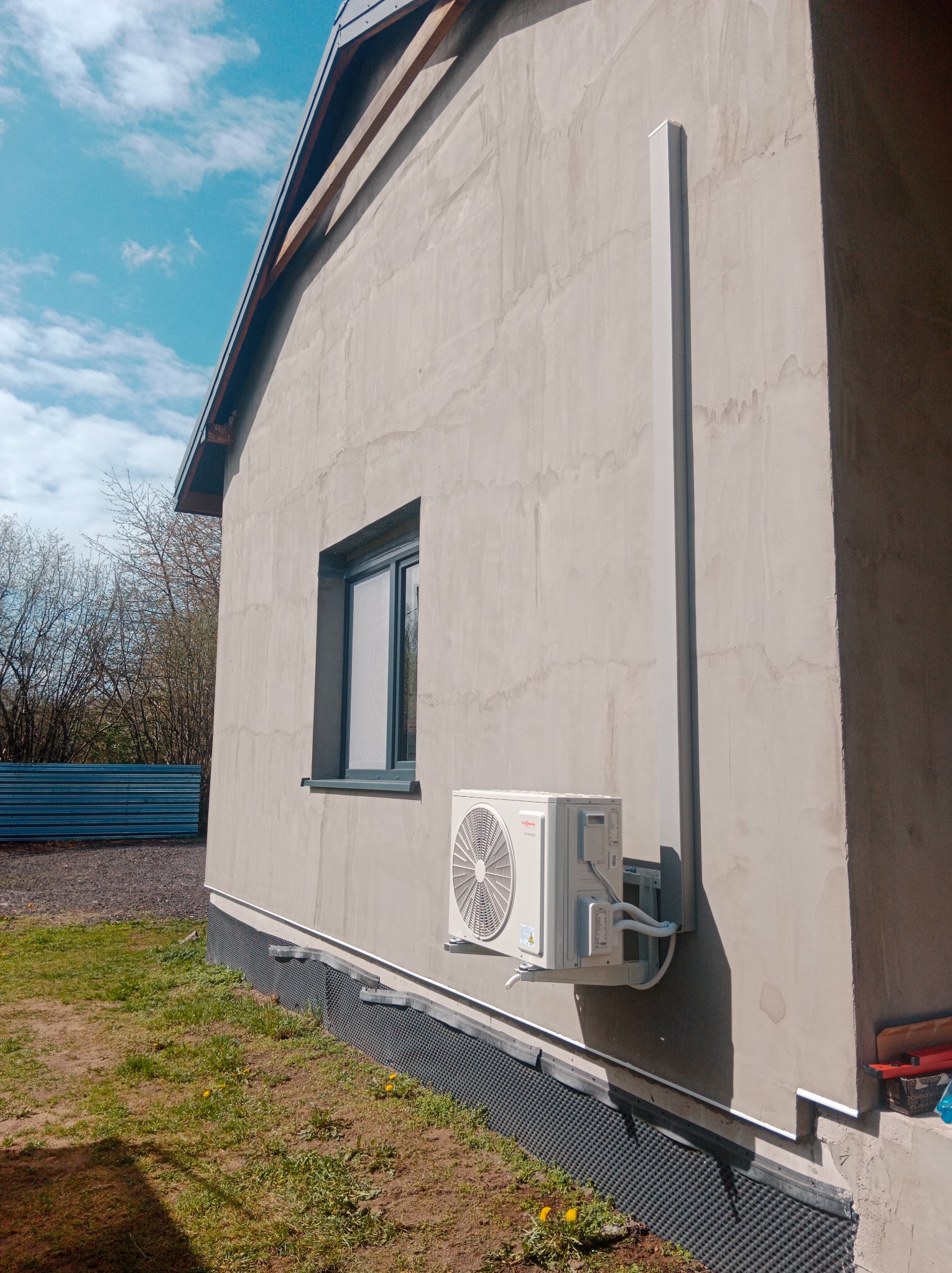 Zewnętrzny montaż klimatyzatora Viessmann na szarej ścianie budynku z widoczną rurą odprowadzającą i fragmentem trawnika z żółtymi kwiatami.