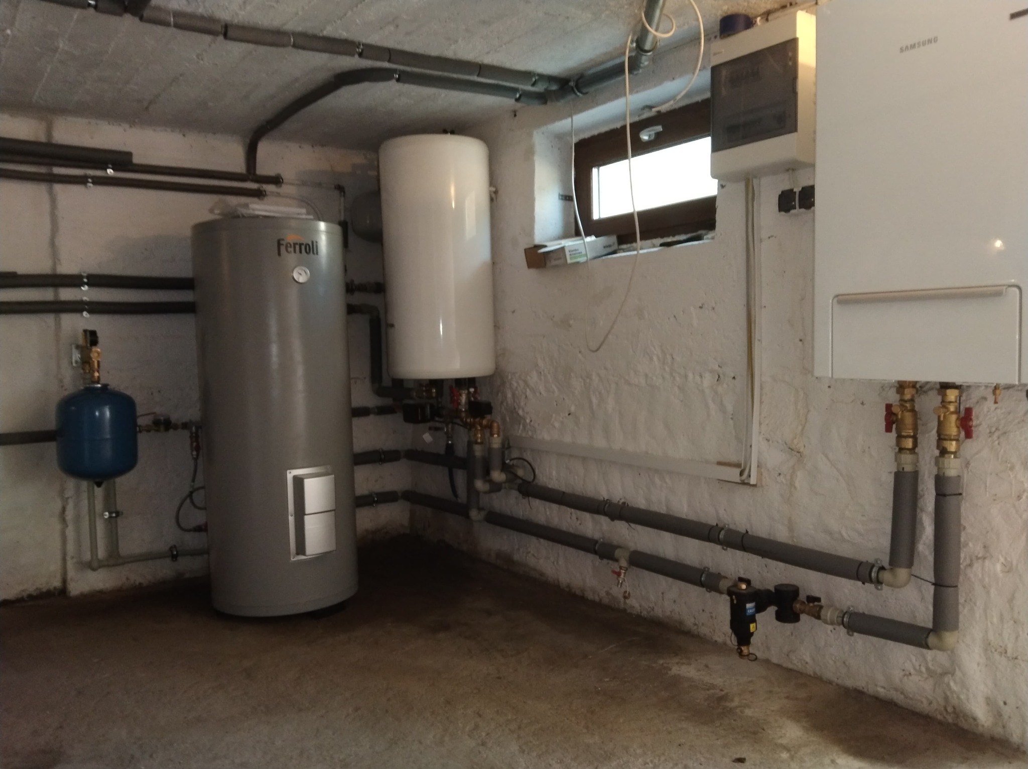 Piwnica z zainstalowanym szarym zbiornikiem Ferroli, białym zasobnikiem ciepłej wody, niebieskim naczyniem przeponowym i białym piecem gazowym Samsung, połączonymi rurami.