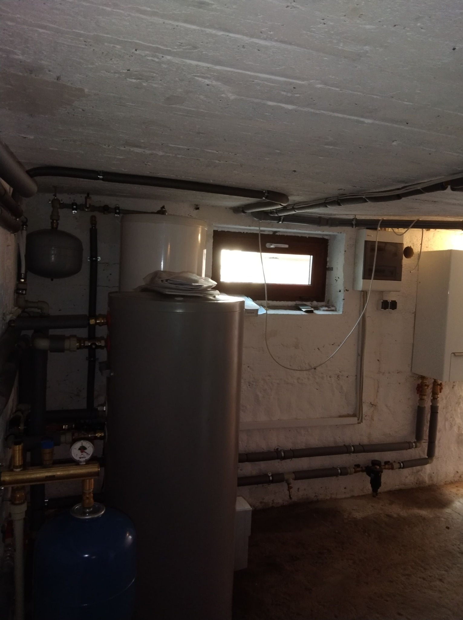 Kotłownia w piwnicy z białymi ścianami, widoczne zbiorniki, rury, okno i bojler.