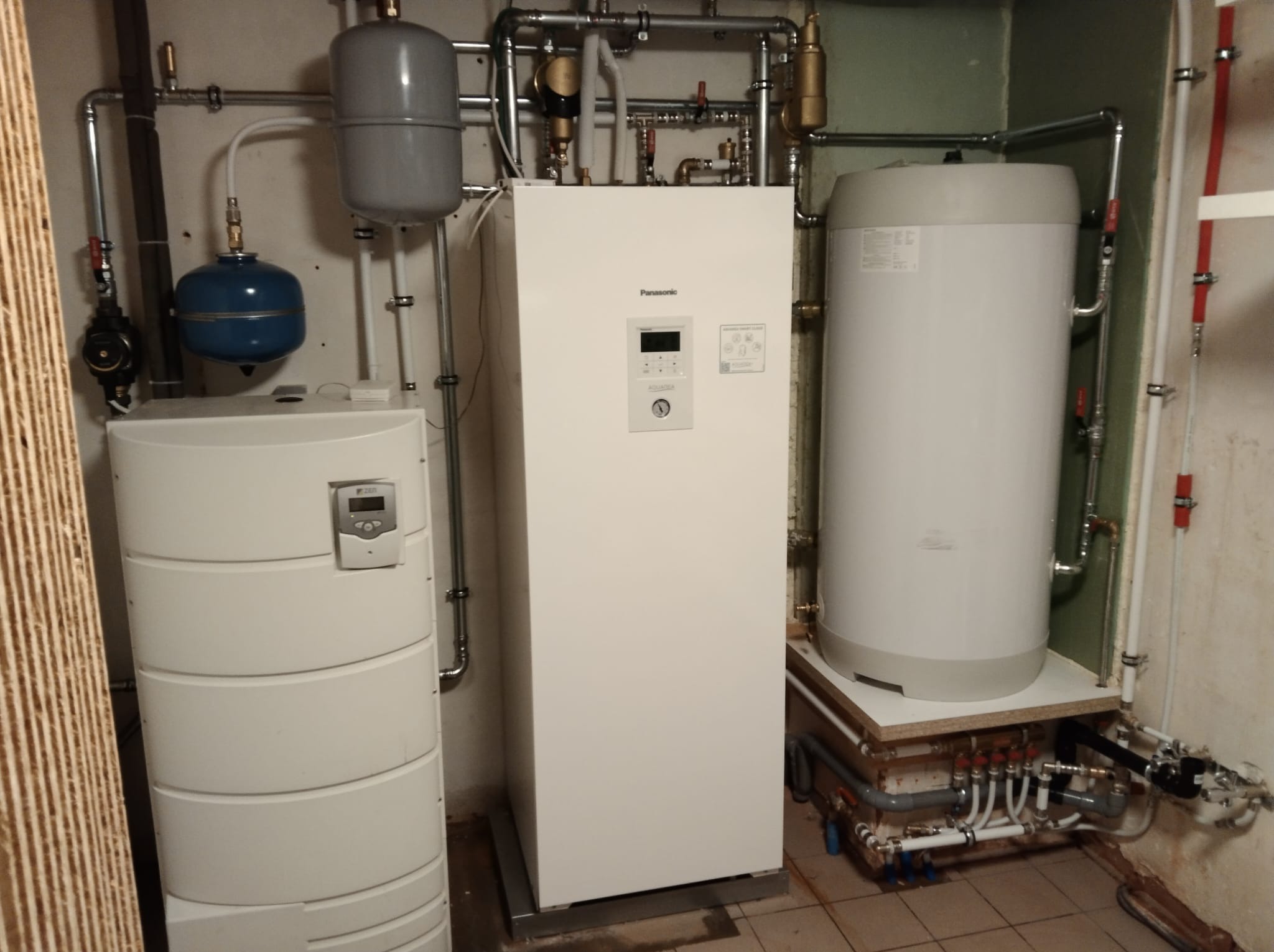 Biała pompa ciepła Panasonic Aquarea z podłączonymi rurami instalacji grzewczej, zbiornik buforowy i naczynie wzbiorcze w kotłowni.