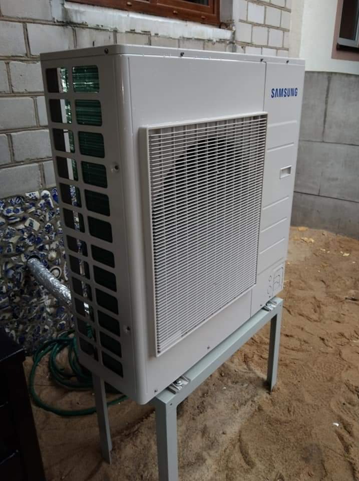 Zewnętrzna jednostka rekuperacji Samsung na stalowym stelażu, zamontowana przy ścianie budynku z dekoracyjnym elementem mozaiki ceramicznej i widocznym oknem.