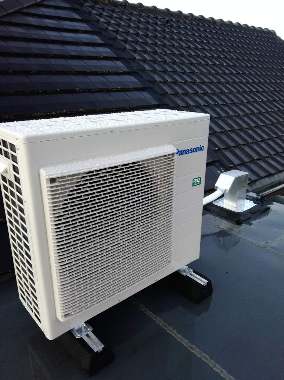 Zewnętrzna jednostka klimatyzacji Panasonic R32 zamontowana na dachu pokrytym ciemną dachówką, widoczne czarne podkładki antywibracyjne i srebrny komin wentylacyjny.