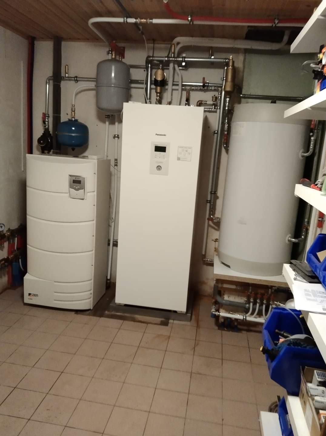 Kotłownia z dwoma białymi piecami, bojlerem i rozbudowaną instalacją hydrauliczną z rur miedzianych i plastikowych, widoczne także zbiorniki wyrównawcze.