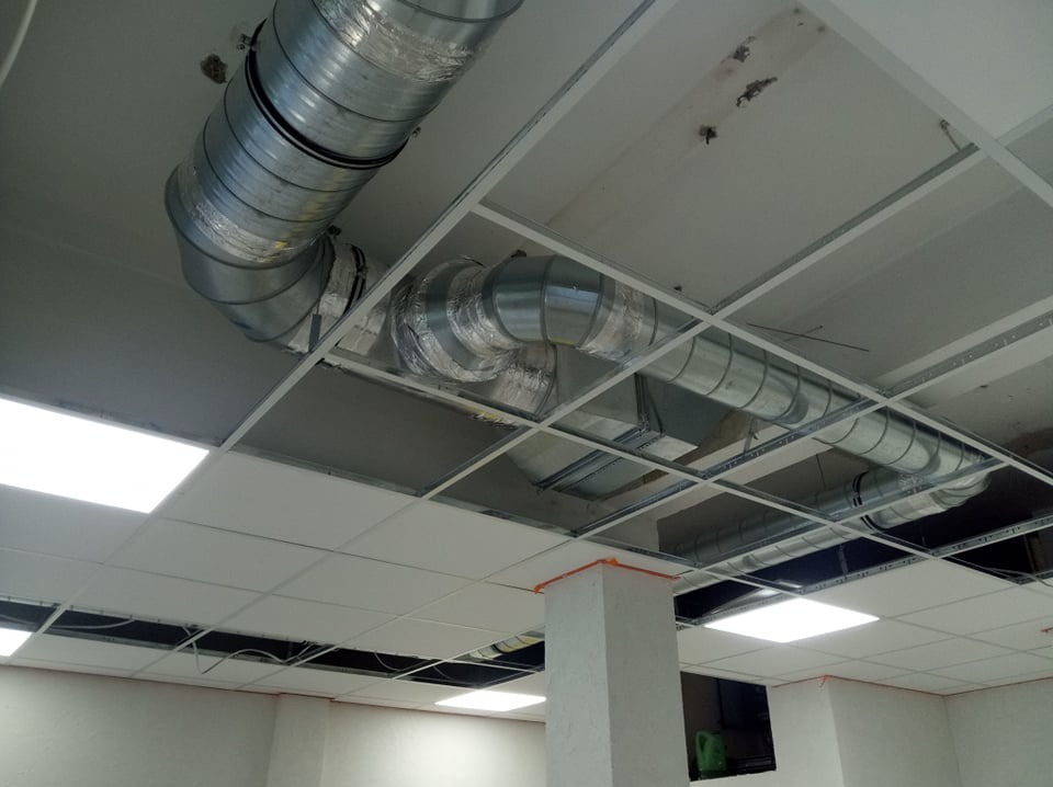 Widok na sufit podwieszany z widocznymi elementami instalacji wentylacyjnej: metalowe rury z izolacją, kratownica sufitowa i panele oświetleniowe.