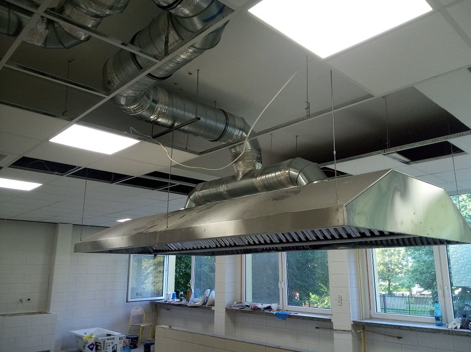 Duża, metalowa, przemysłowa okap kuchenny zawieszony pod sufitem z widocznymi rurami wentylacyjnymi w pomieszczeniu z białymi kafelkami i oknami.