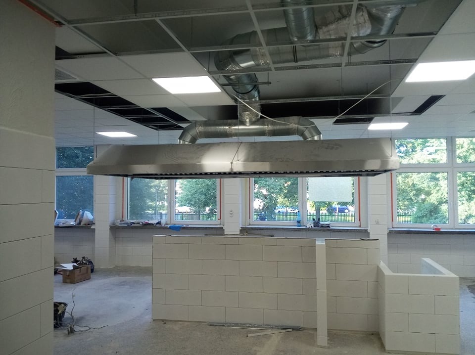 Wnętrze komercyjne w trakcie wykańczania: widoczne ściany z białych bloczków, okna z widokiem na zieleń, oraz system wentylacji z metalową osłoną nad przyszłym stanowiskiem pracy. Na suficie...