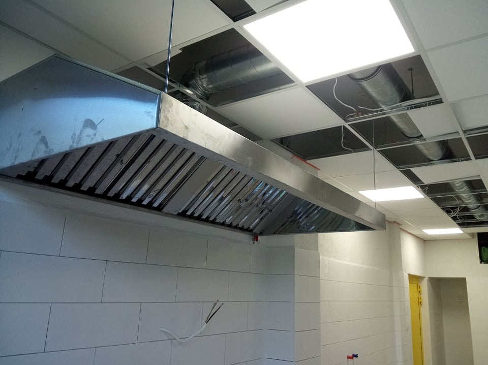Długa, metalowa okap kuchenny zawieszony nad białą ścianą z płytek w pomieszczeniu z podwieszanym sufitem i widocznymi rurami wentylacyjnymi.