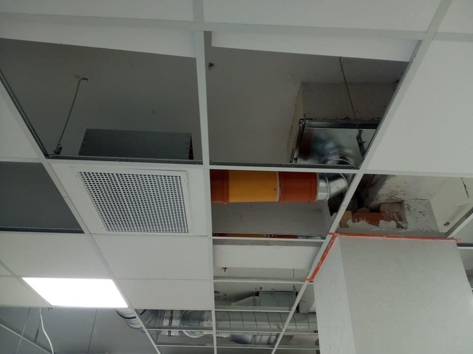 Sufit podwieszany z widoczną instalacją wentylacyjną: pomarańczowo-żółty kanał wentylacyjny, kratka wentylacyjna oraz fragmenty instalacji ukryte za płytami sufitowymi.