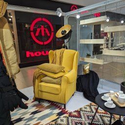 MeblePasternak - Żółty fotel z poduszkami i pledem na wzorzystym dywanie, obok stolik z filiżankami, w tle ekspozycja sklepu odzieżowego.